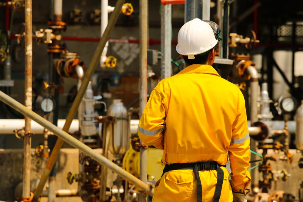 Back of a field operator wearing yellow coverall with petrochemical plant in the background.